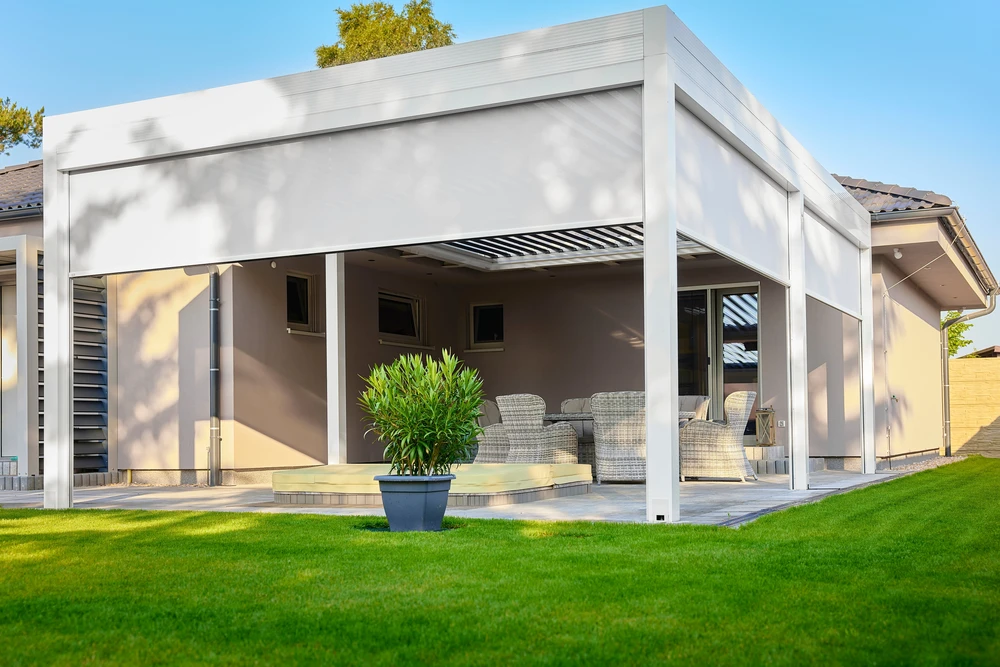 Une pergola fixe en aluminium gris clair ou anthracite installée sur une terrasse résidentielle moderne, créant un espace ombragé et aménagé. La structure affiche des lames ou une toiture louvrable en aluminium, intégrée harmonieusement à l'architecture de la maison. La terrasse est meublée simplement (salon de jardin, chaises) suggérant un usage quotidien comme pièce à vivre extérieure. Ambiance ensoleillée avec ombres nettes créées par la structure. Pas de personnes visibles, pas de texte ou logo.
