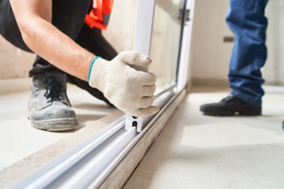 Deux artisans menuisiers professionnels travaillant ensemble sur un chantier extérieur, installant une fenêtre ou une porte en aluminium sur une façade résidentielle. L'un guide et vérifie l'installation tandis que l'autre intervient. L'image montre clairement leur expertise commune, leur coordination professionnelle et leur présence simultanée sur le chantier, illustrant le duo de menuiserie de Fermeture Pro Sage.
