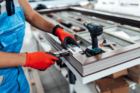 Artisan menuisier en tenue professionnelle discutant avec un client devant une fenêtre ou porte d'entrée à son domicile, montrant plans ou documents techniques, illustrant le diagnostic et le suivi personnalisé du projet de fermetures.