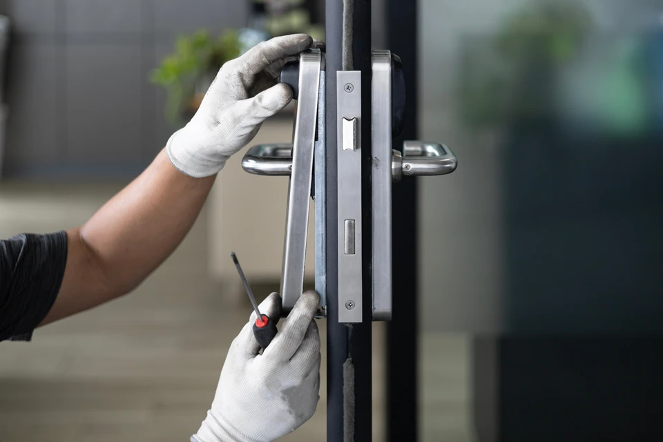 Un artisan en t-shirt bleu installe une serrure sur une porte d'entrée blanche dans un intérieur résidentiel