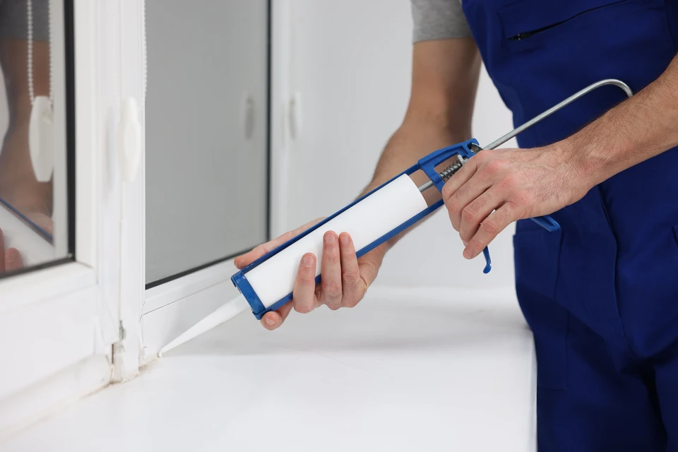 Ouvrier en bâtiment portant casque de sécurité blanc et salopette bleue utilise une perceuse électrique sur cadre de fenêtre dans espace intérieur moderne avec baies vitrées